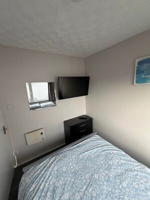 A bedroom with a bed, a flat-screen TV mounted on the wall, a window with blinds, a radiator, and a small bedside table with a remote control. - Chalet 91A South Shore Bridlington, Sleeps 4 (Bridlington)