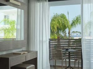 A modern room with a large window, a wooden desk, a chair, and a balcony with a view of palm trees and the ocean. - Atlantica Sancta Napa Resort (Ayia Napa)