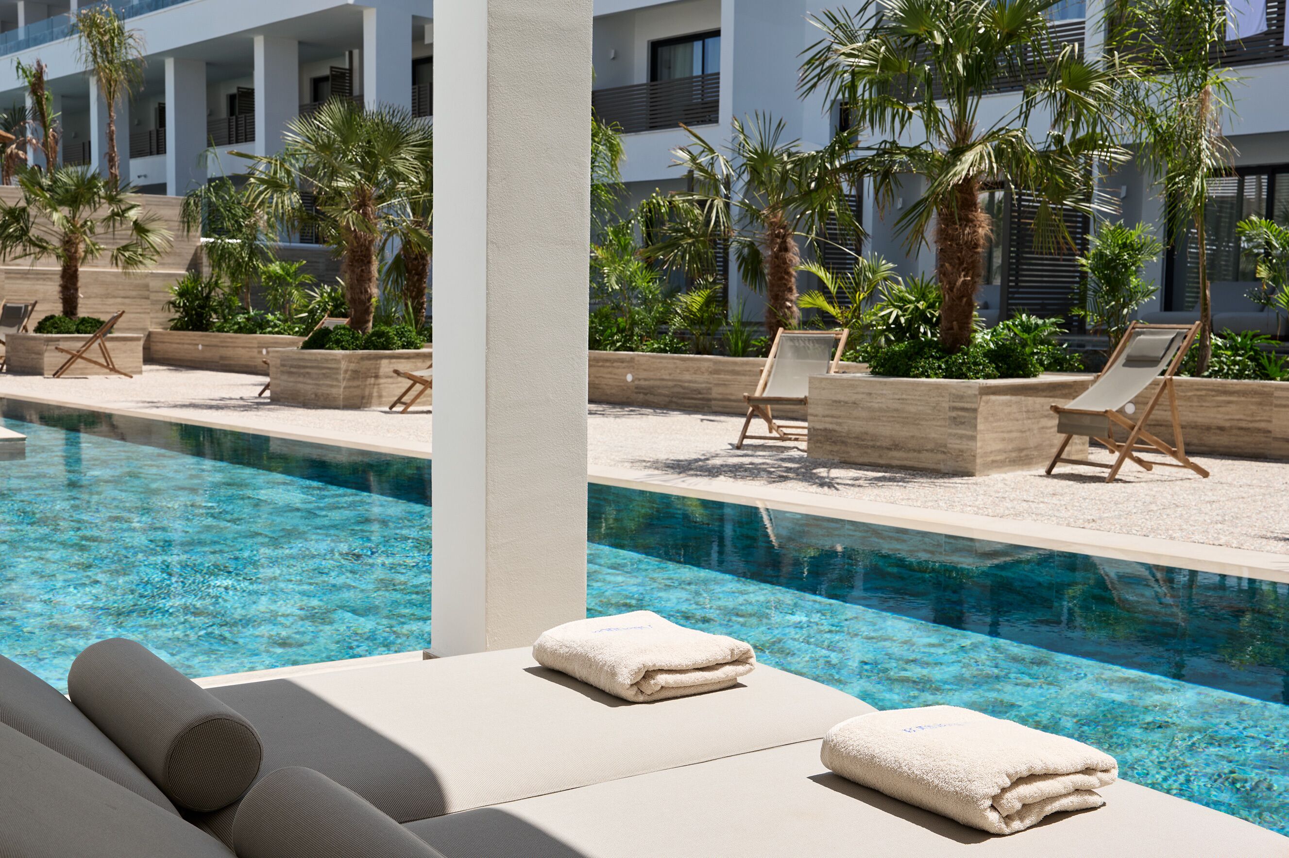 A poolside area with lounge chairs and towels, surrounded by palm trees and a modern building.
