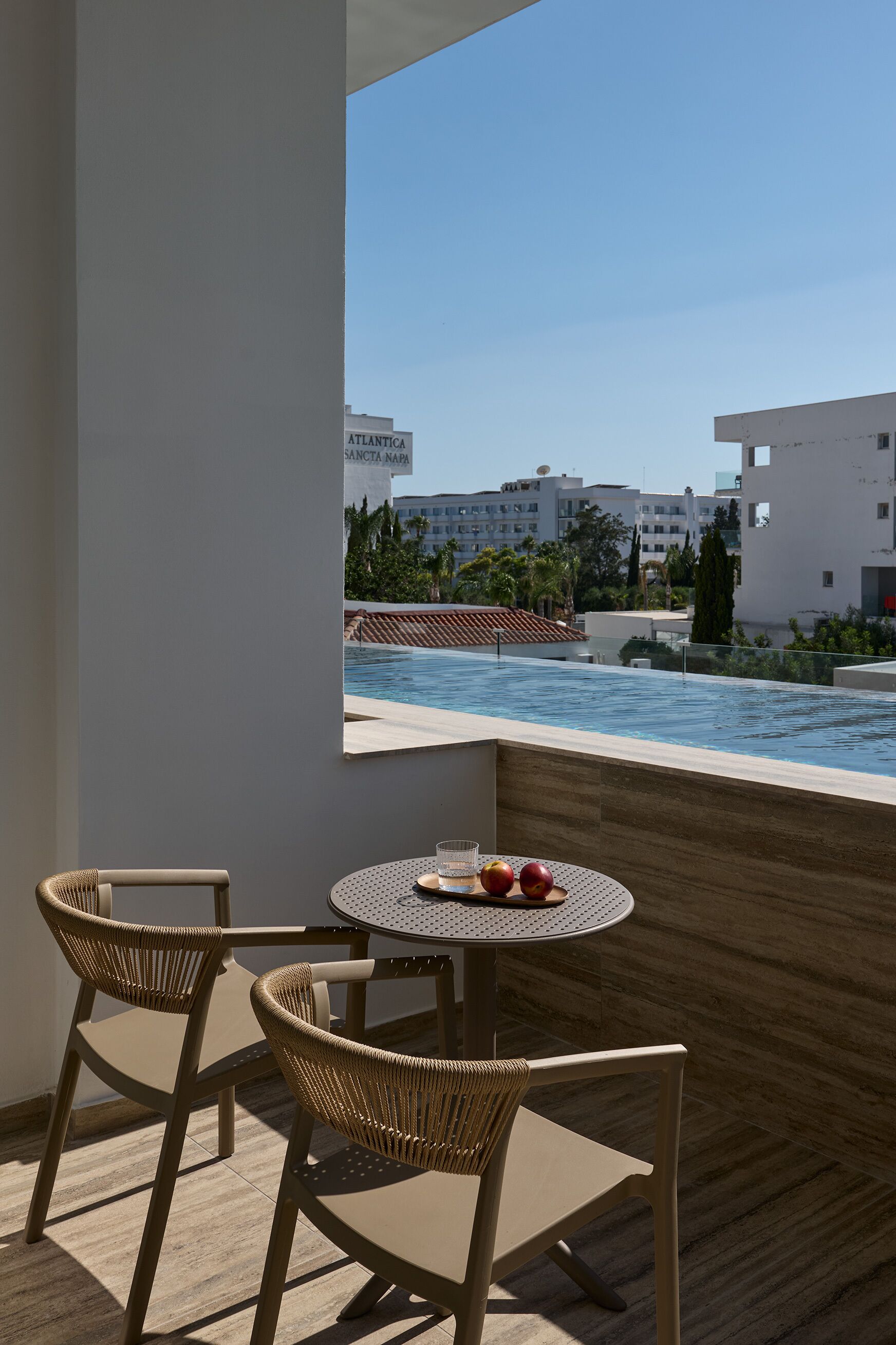 Un balcon avec deux fauteuils en osier et une petite table ronde, donnant sur une piscine et sur des bâtiments en arrière-plan.