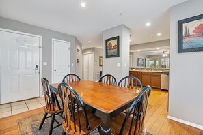 A dining area with a wooden table and chairs, a kitchen in the background, and a hallway with doors.