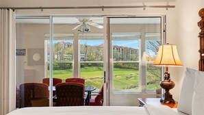 Habitación de hotel con cama, una mesa con sillas, una lámpara y vista a un campo de golf a través de amplias ventanas.