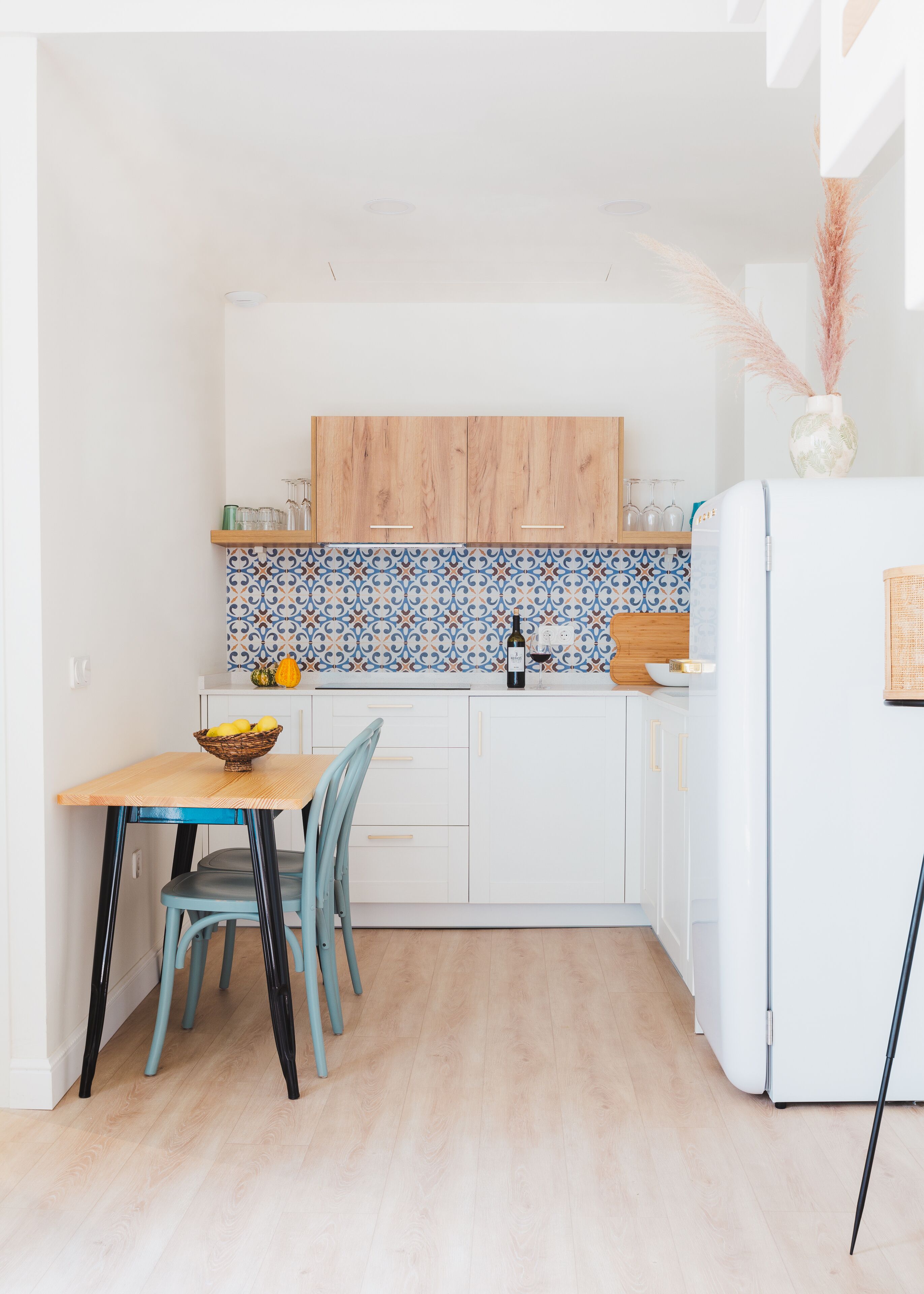 Une cuisine de taille réduite avec des meubles blancs, un crédence bleue à motifs, un plan de travail en bois et un réfrigérateur blanc.