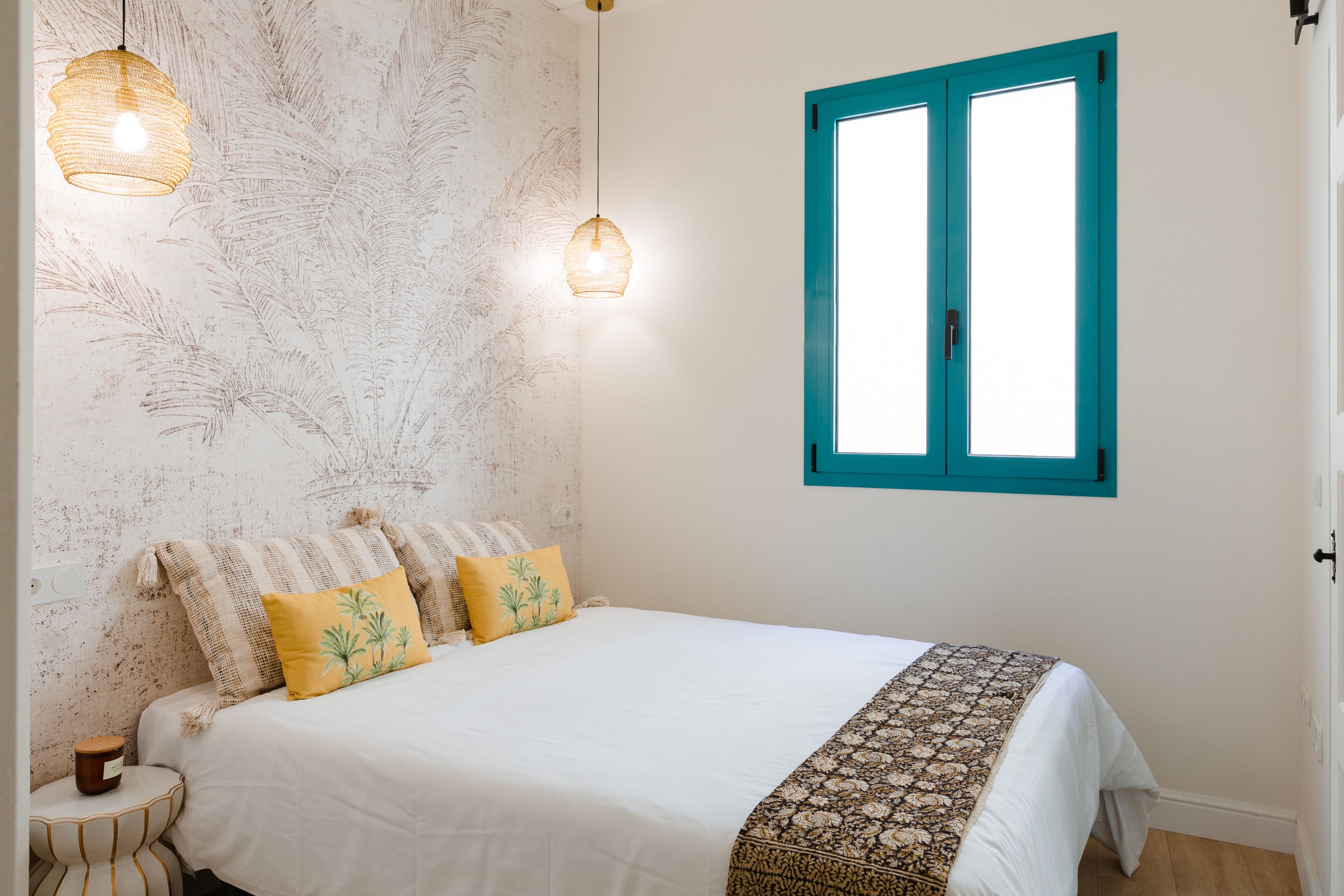 A bedroom with a bed, two hanging lamps, a window with blue shutters, and a patterned wall.