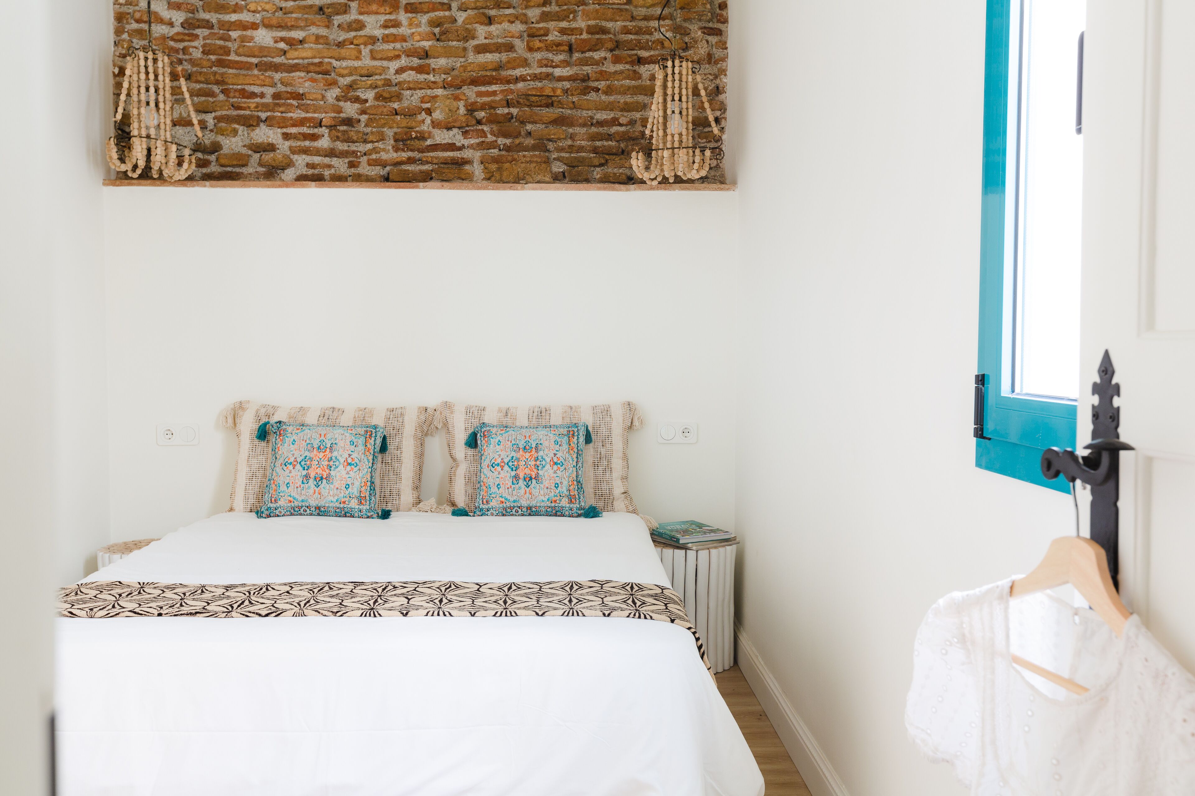Un dormitorio con pared de ladrillo, cama con ropa de cama estampada, mesita de noche pequeña y ventana con marco azul.