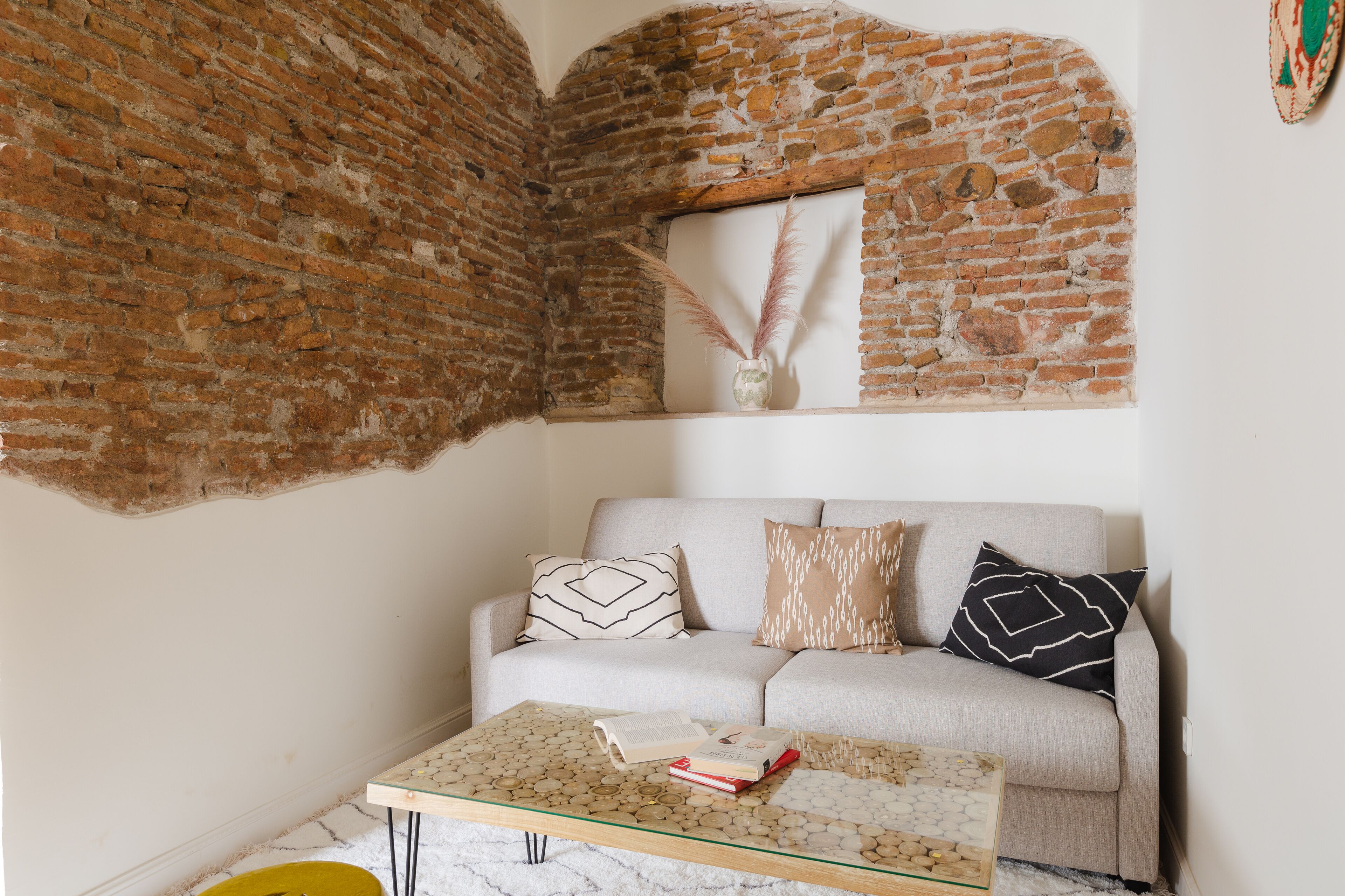 A living room with a brick wall, a sofa with patterned cushions, and a glass coffee table with books.