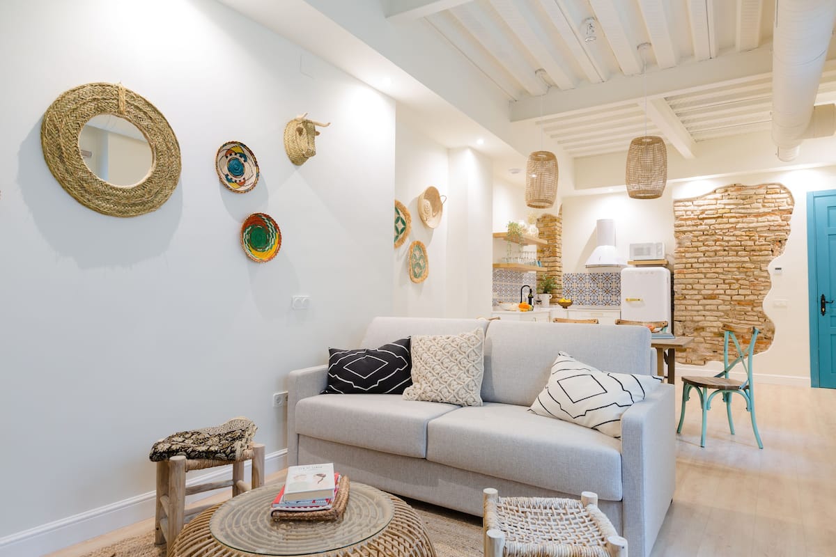 Una sala moderna con un sofá de color gris claro, una mesa de centro redonda y una silla de mimbre. Objetos decorativos tejidos se exhiben en las paredes.