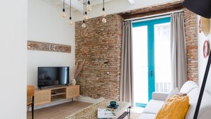 Un salon avec un mur en briques, un téléviseur posé sur un meuble en bois, une table basse en verre, un canapé agrémenté d’un coussin jaune et une porte avec des moulures de couleur turquoise.