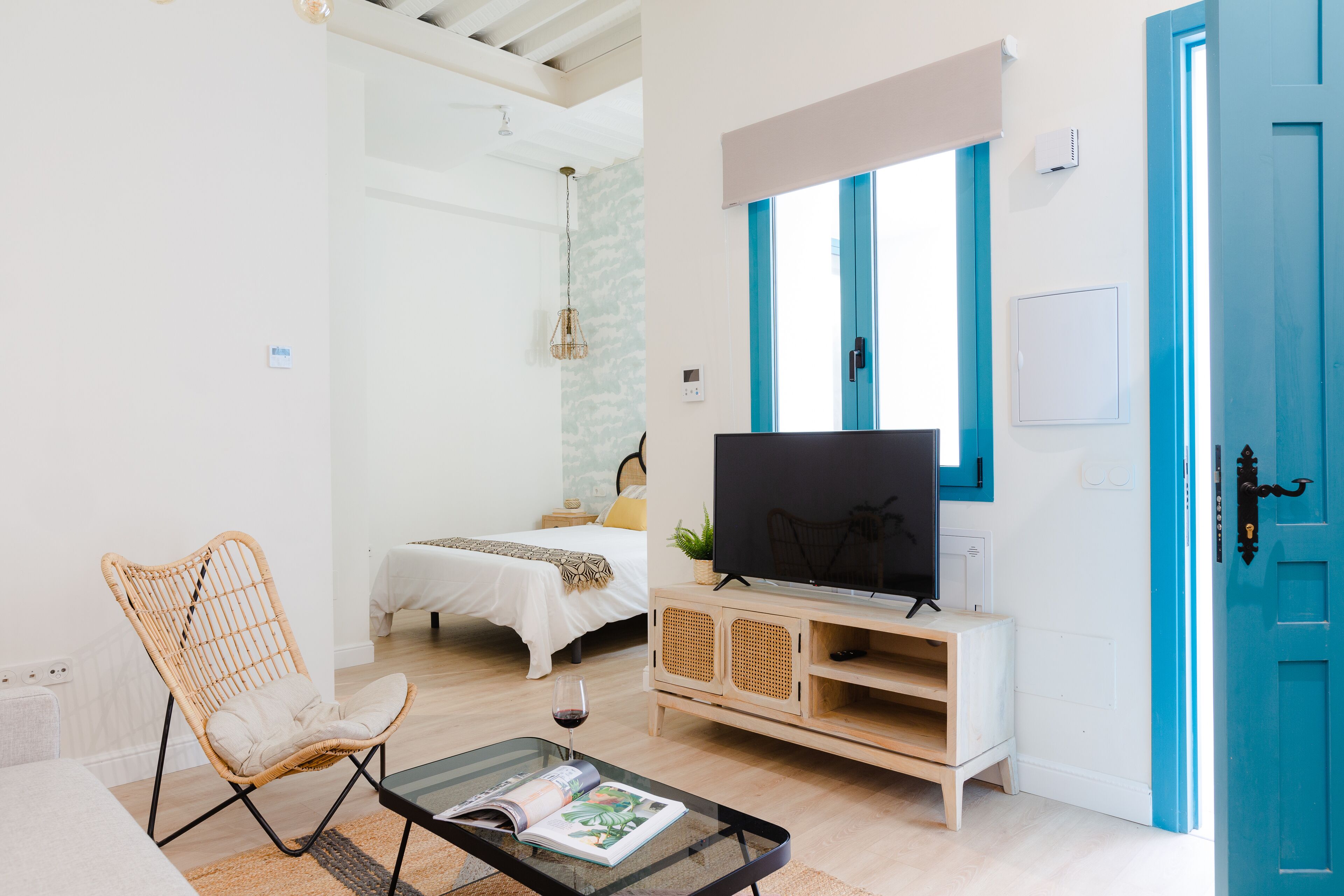 A modern living room with a glass coffee table, a wicker chair, a bed with a white duvet, a television on a wooden cabinet, and a blue door.
