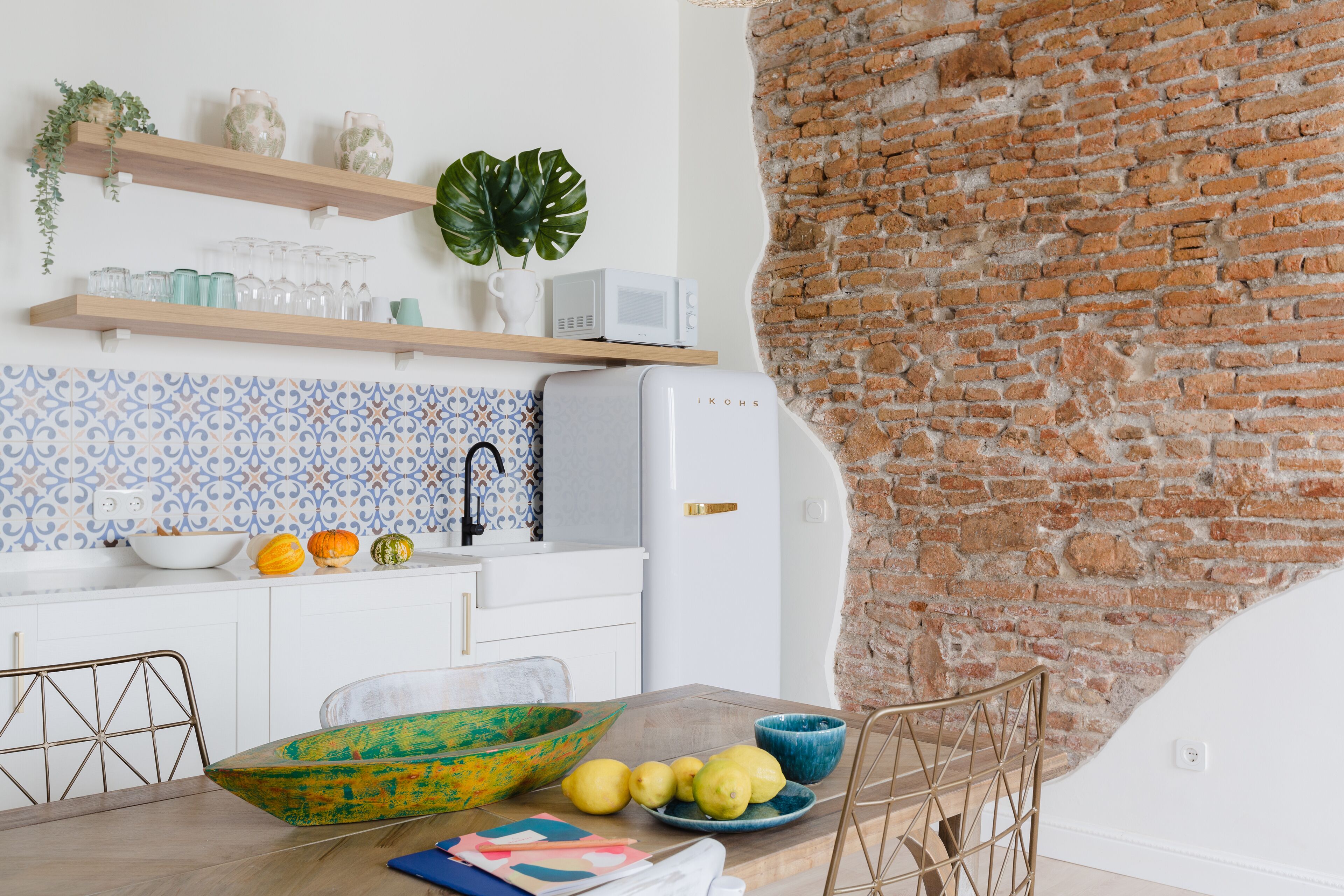 A kitchen with white cabinets, a white refrigerator, a microwave, and a brick wall. There are shelves with glassware and a plant.