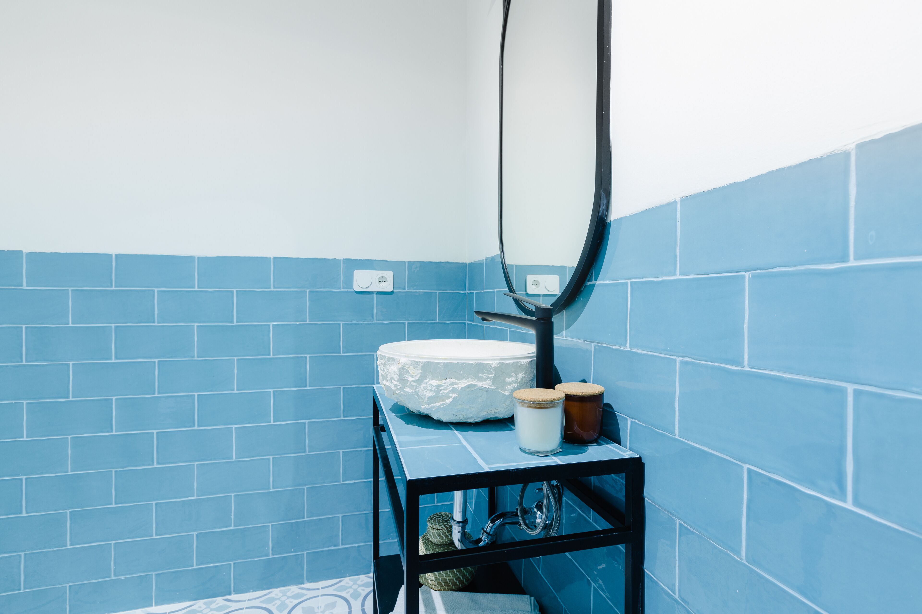 Bagno con piastrelle blu, un lavandino bianco, uno specchio e uno scaffale nero con un vasetto e una candela.