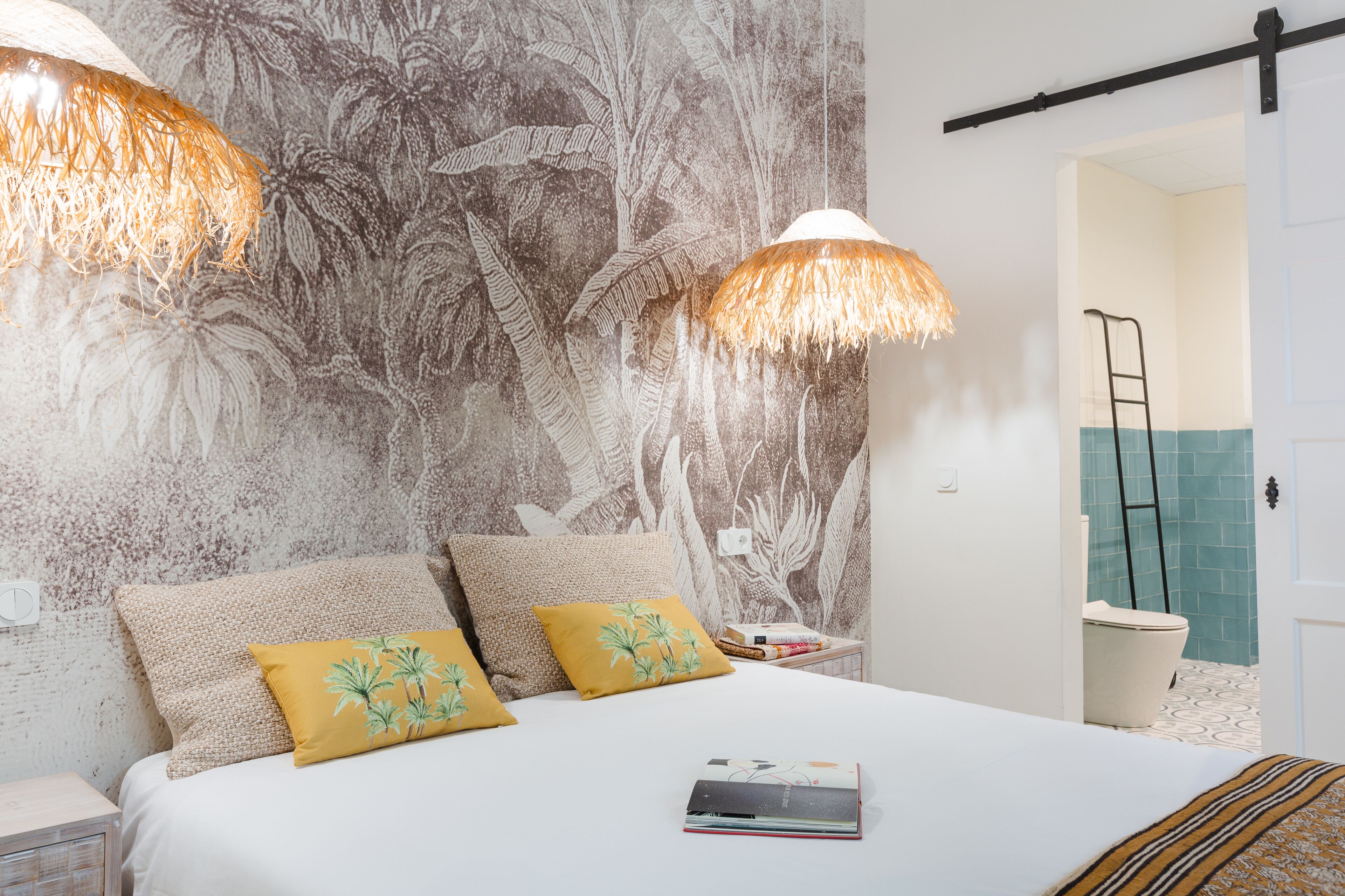 A bedroom with a bed, two pillows, a nightstand, a wall with a leaf pattern, and a door leading to a bathroom.
