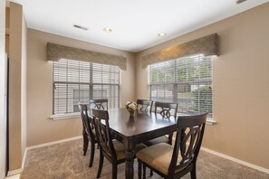 A dining room with a dark wooden table and chairs, beige walls, and large windows with blinds.