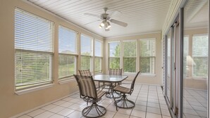 Un solárium con una mesa de comedor y sillas, ventanas amplias, un ventilador de techo y vistas al exterior con vegetación.