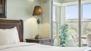 A neatly made bed with white linens, a wooden nightstand with a lamp, a potted plant, and a chair by a window with a view of trees. - Walk-In w/2 King Bd/ 2 Ba Golf Course/ Deals on Golf/Tickets! (Branson)