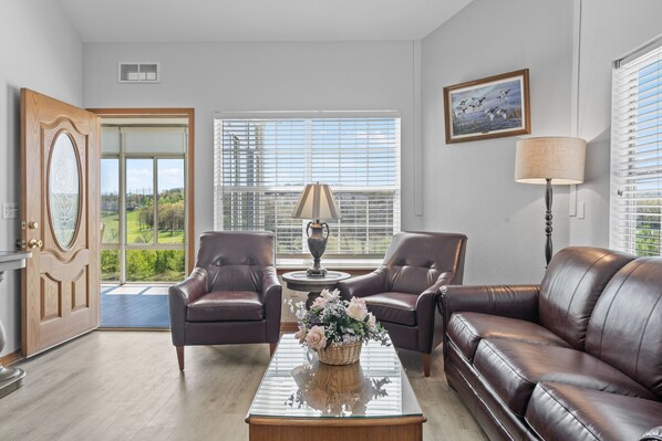A living room with a brown leather sofa, two armchairs, a glass coffee table with a floral centerpiece, a lamp, and a view of greenery through large windows.