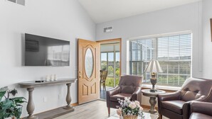 A living room with a flat-screen TV mounted on the wall, a wooden door, a window with blinds, two leather armchairs, a coffee table with a vase, and a side table with a lamp. - 3 Bedroom, 3 Bath Golf Condo with Beautiful Course View from All-Season Sunroom (Branson)