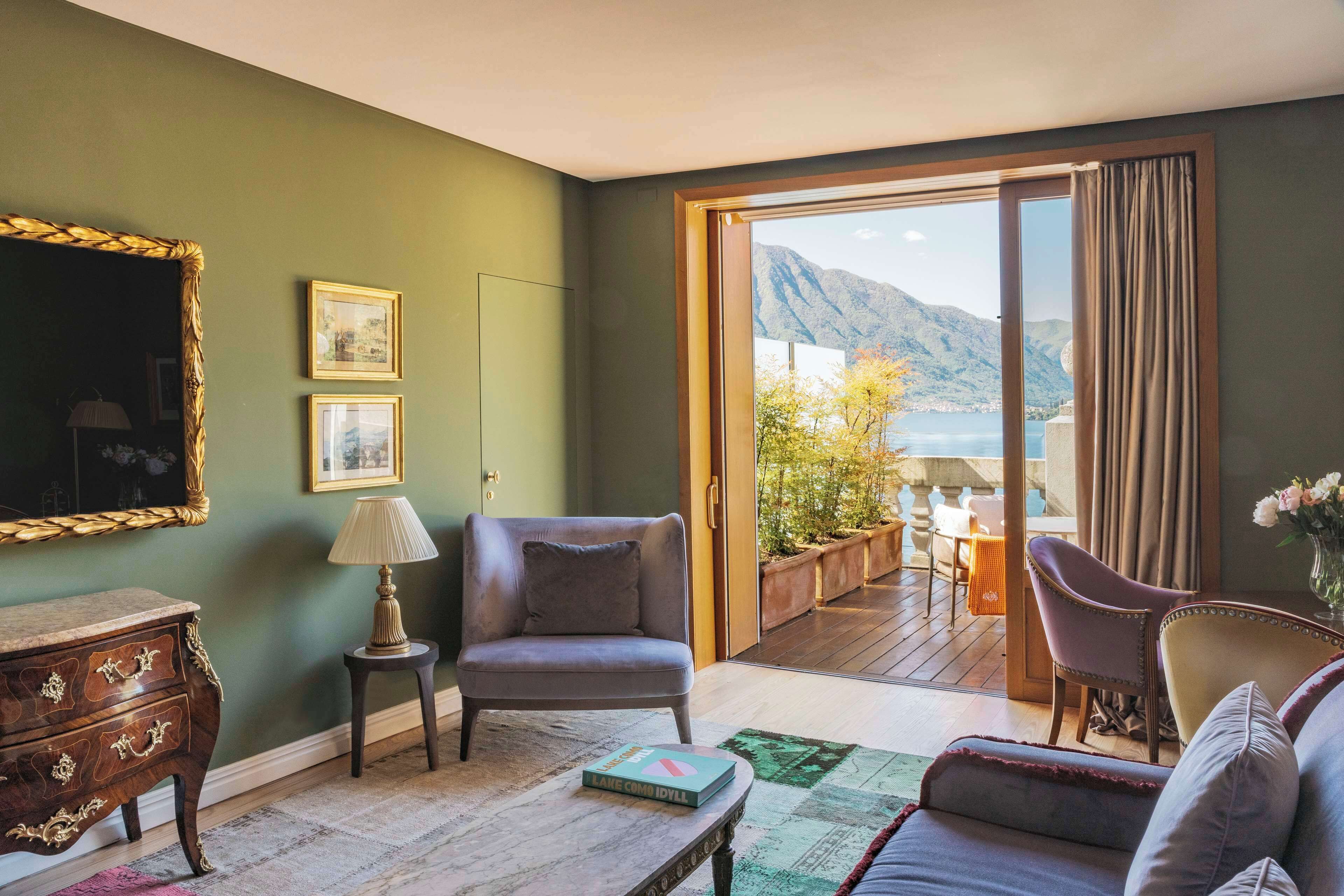 A room with a view of mountains and a lake, a wooden dresser, a grey armchair, a small table with a book, and a sofa.