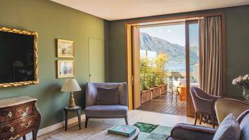 A room with a view of mountains and a lake, a wooden dresser, a grey armchair, a small table with a book, and a sofa.