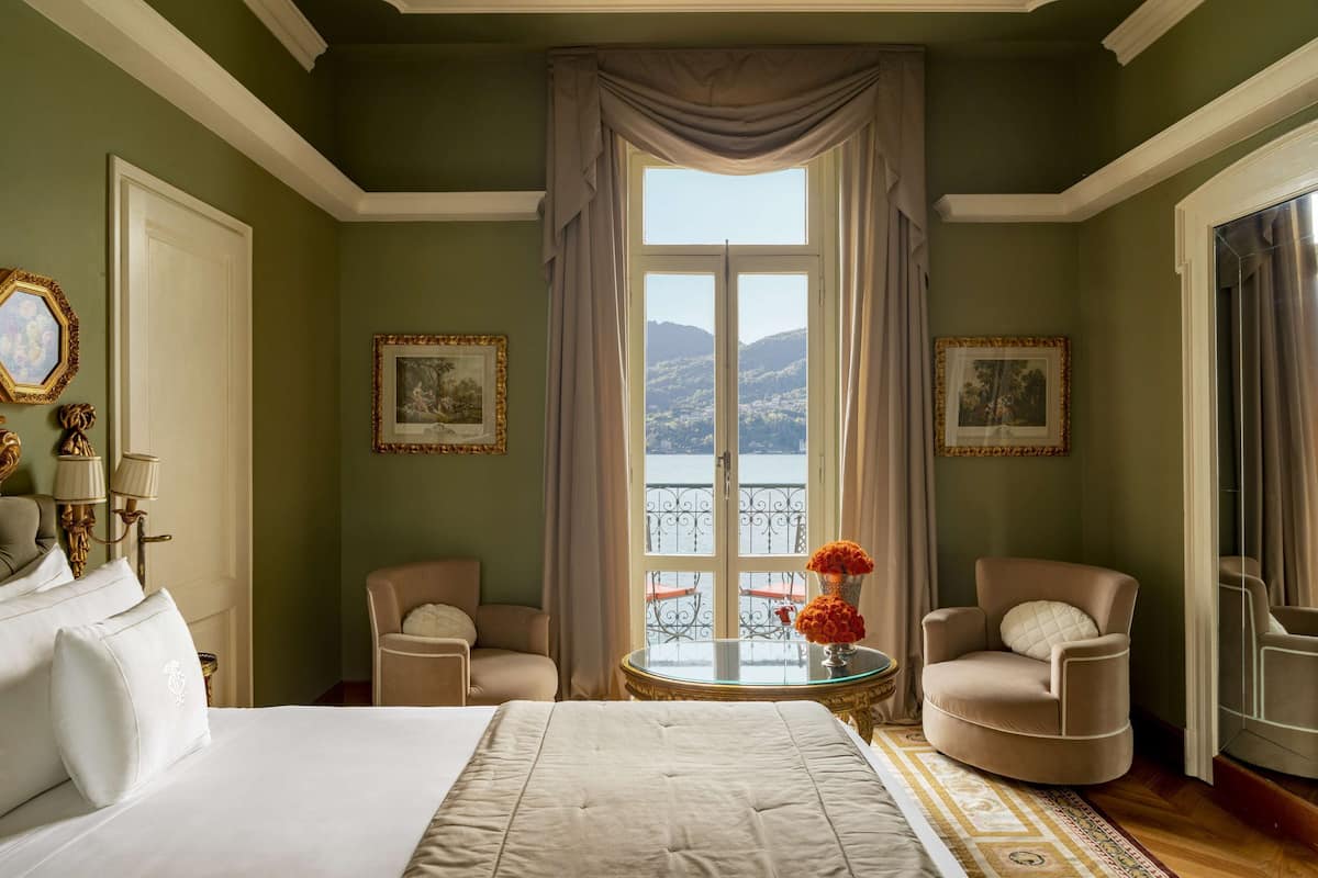 A hotel room with a large bed, two armchairs, a small table with a vase of flowers, and a view of a balcony with a railing.