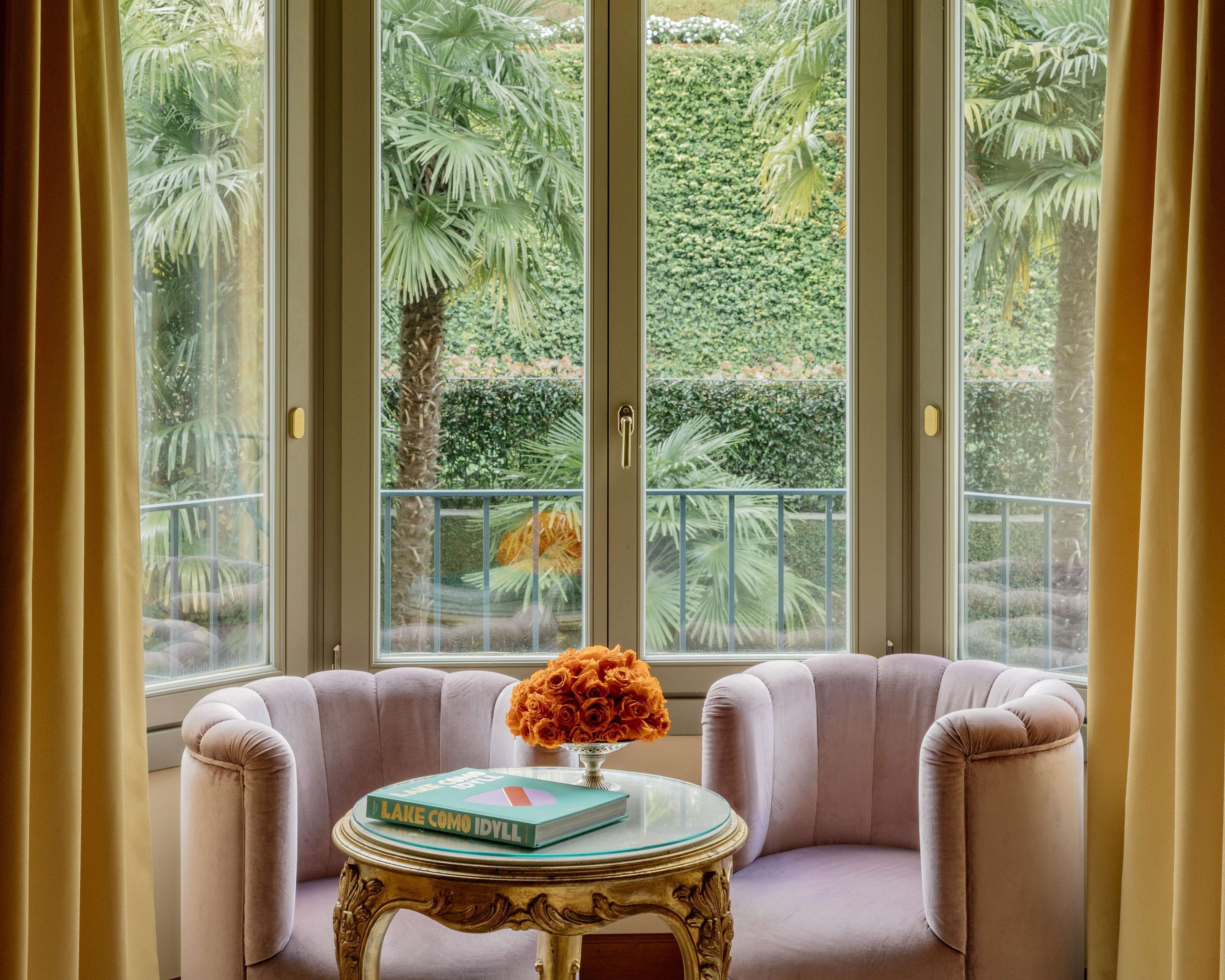 A room with a large window, a plush armchair, a small table with a book and a vase of flowers.
