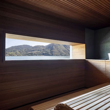 A modern room with a wooden slatted ceiling, a large window overlooking a lake and mountains, and a wooden bench.