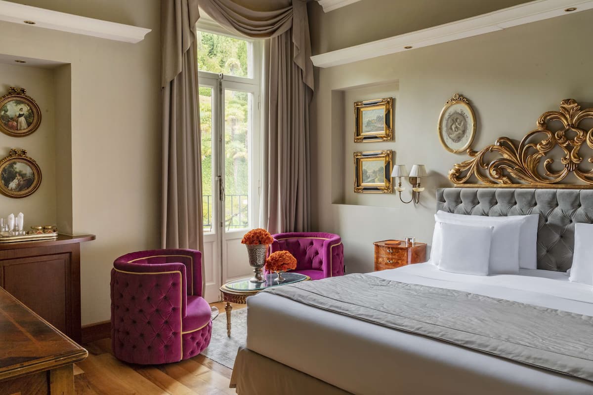 A bedroom with a large bed, a vanity with a mirror, and a chair. There are framed pictures on the wall and a window with curtains.