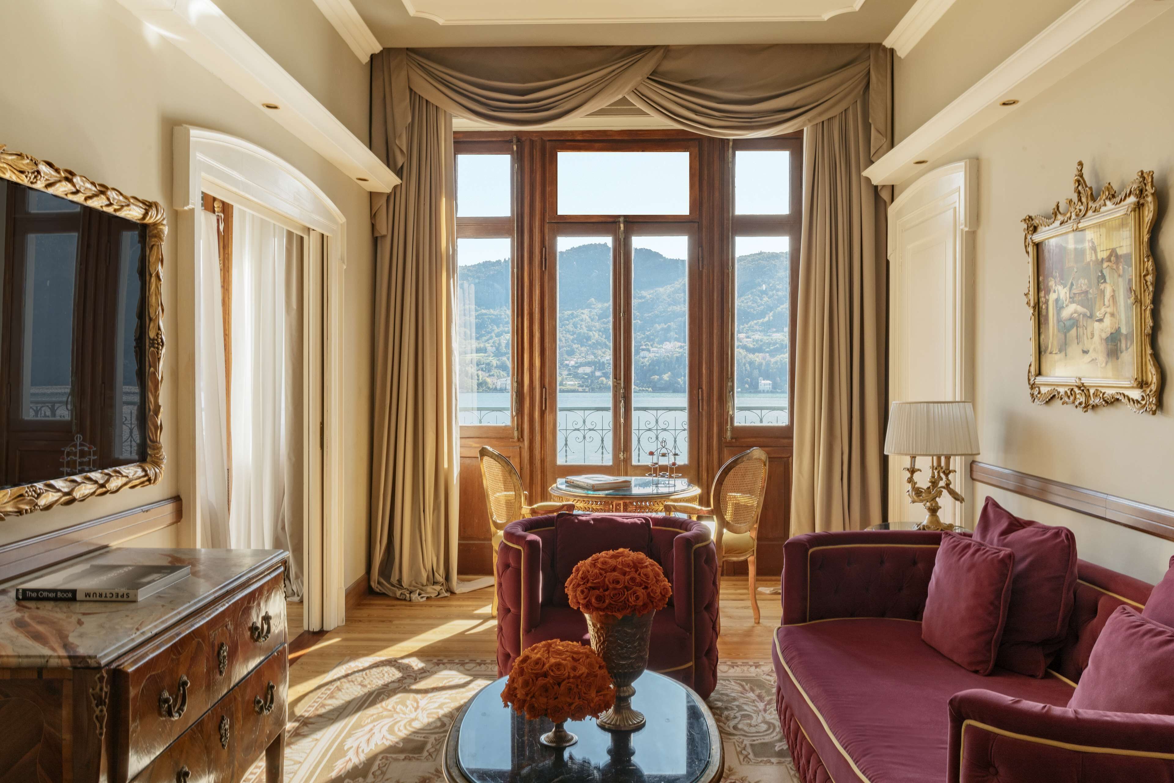Una habitación lujosa con un ventanal grande, un sofá rojo y mullido, un mueble de madera y una mesa con un jarrón de flores.