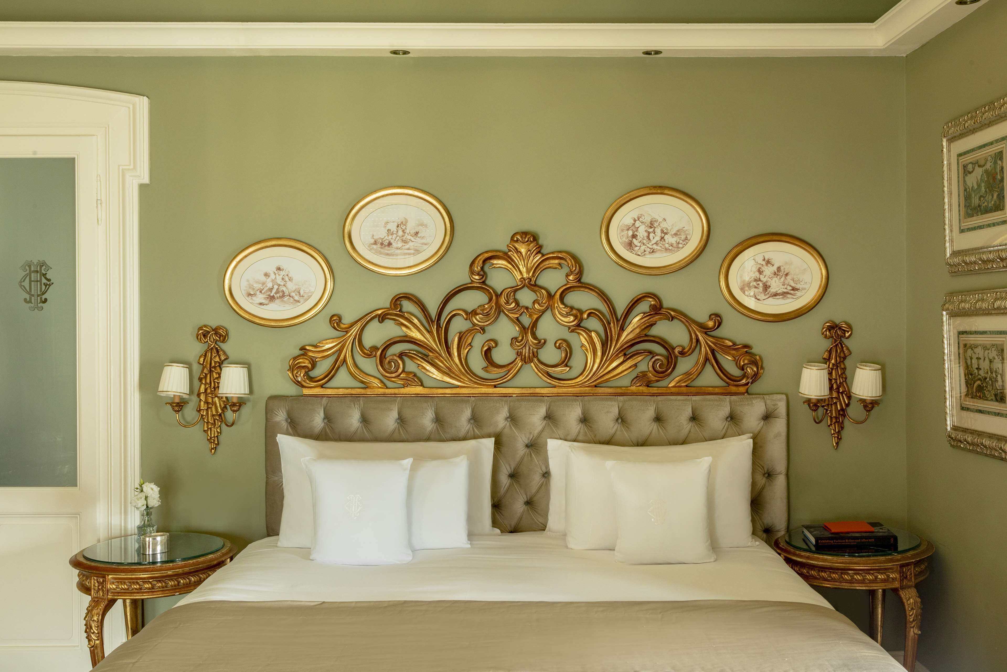 Une chambre à coucher avec une tête de lit capitonnée, quatre tableaux encadrés, deux lampes de chevet et une table ronde sur laquelle est posé un vase.