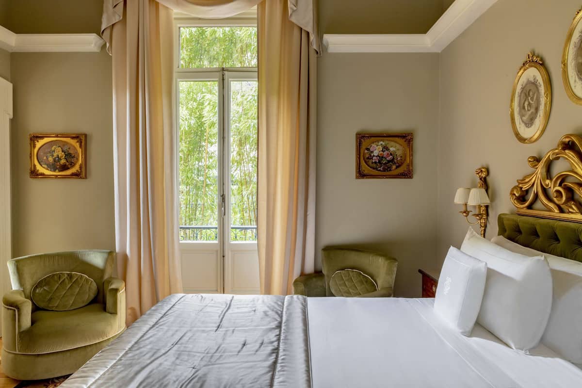 A hotel room with a bed, two chairs, a window with curtains, and wall-mounted lamps.