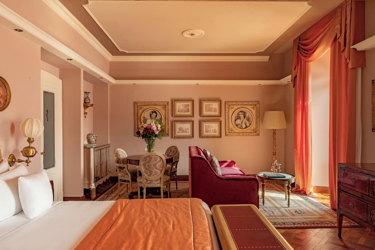 A hotel room with a large bed, a red armchair, a dining table with chairs, and a small side table with a lamp.