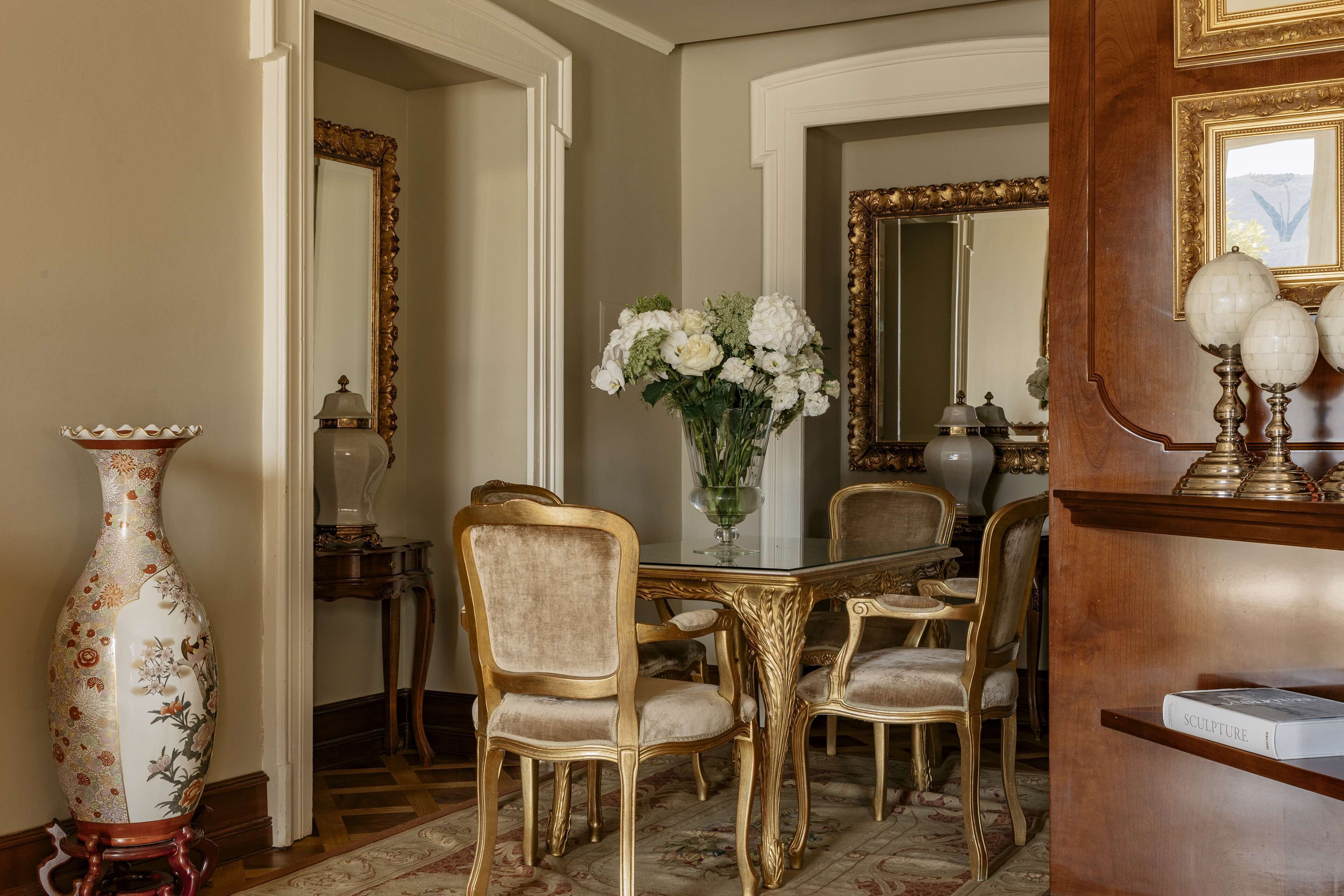 Un comedor elegante con una mesa de vidrio, sillas doradas y un jarrón grande con flores blancas.