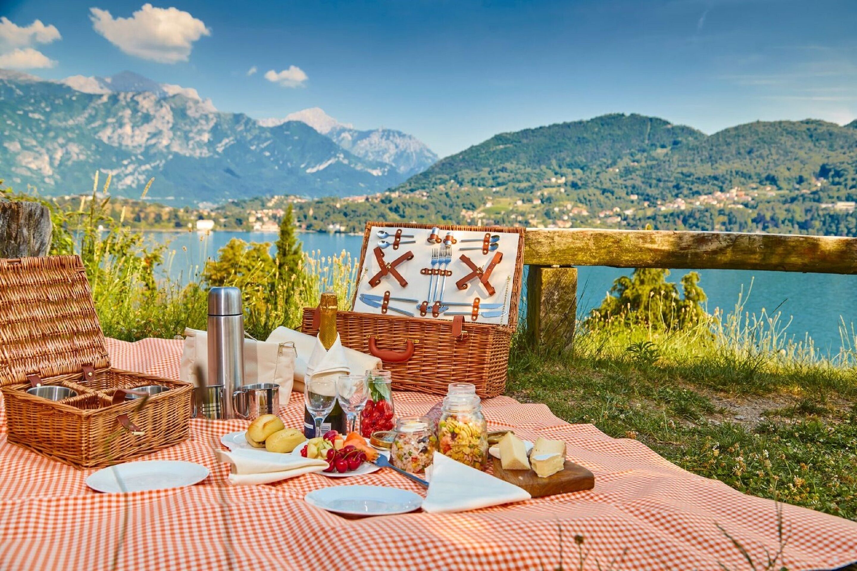 A picnic setup with a wicker basket, a thermos, a bottle, and a picnic table with food items and a signboard, overlooking a lake and mountains.