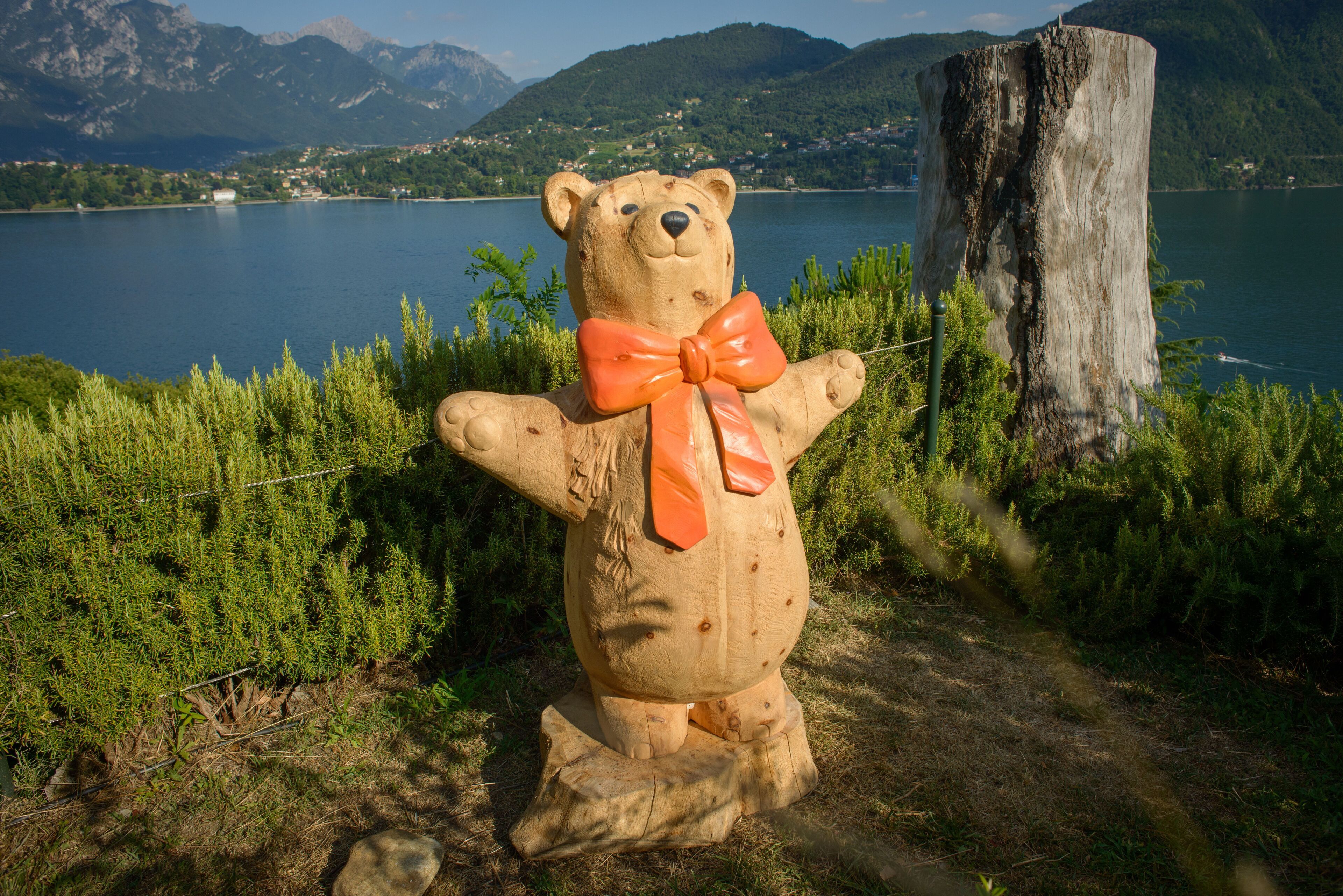 Una escultura de madera de un oso con lazo naranja, frente a un paisaje de lago y montañas.