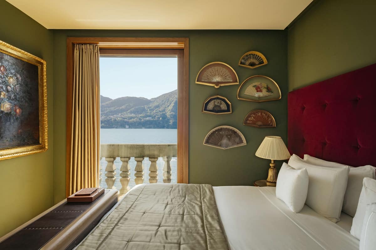 A hotel room with a bed, a nightstand with a lamp, a window with curtains, and a view of a lake and mountains.