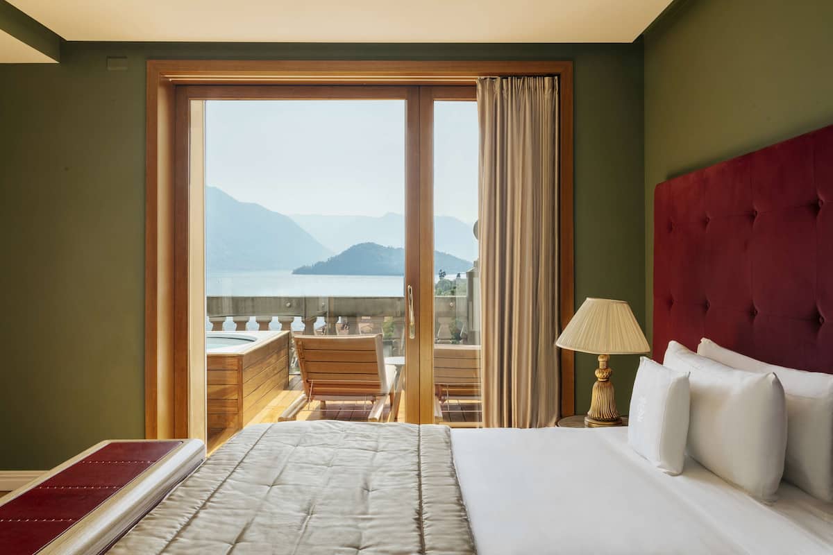 A hotel room with a large bed, a red headboard, a view of a balcony with a wooden chair, and a lamp on the nightstand.