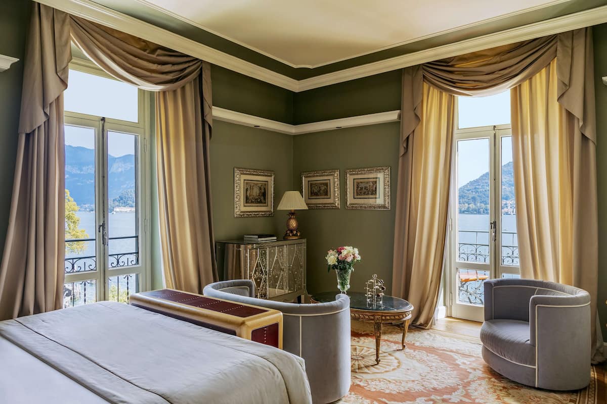 A hotel room with a bed, two armchairs, a small table, and a view of a lake through large windows.