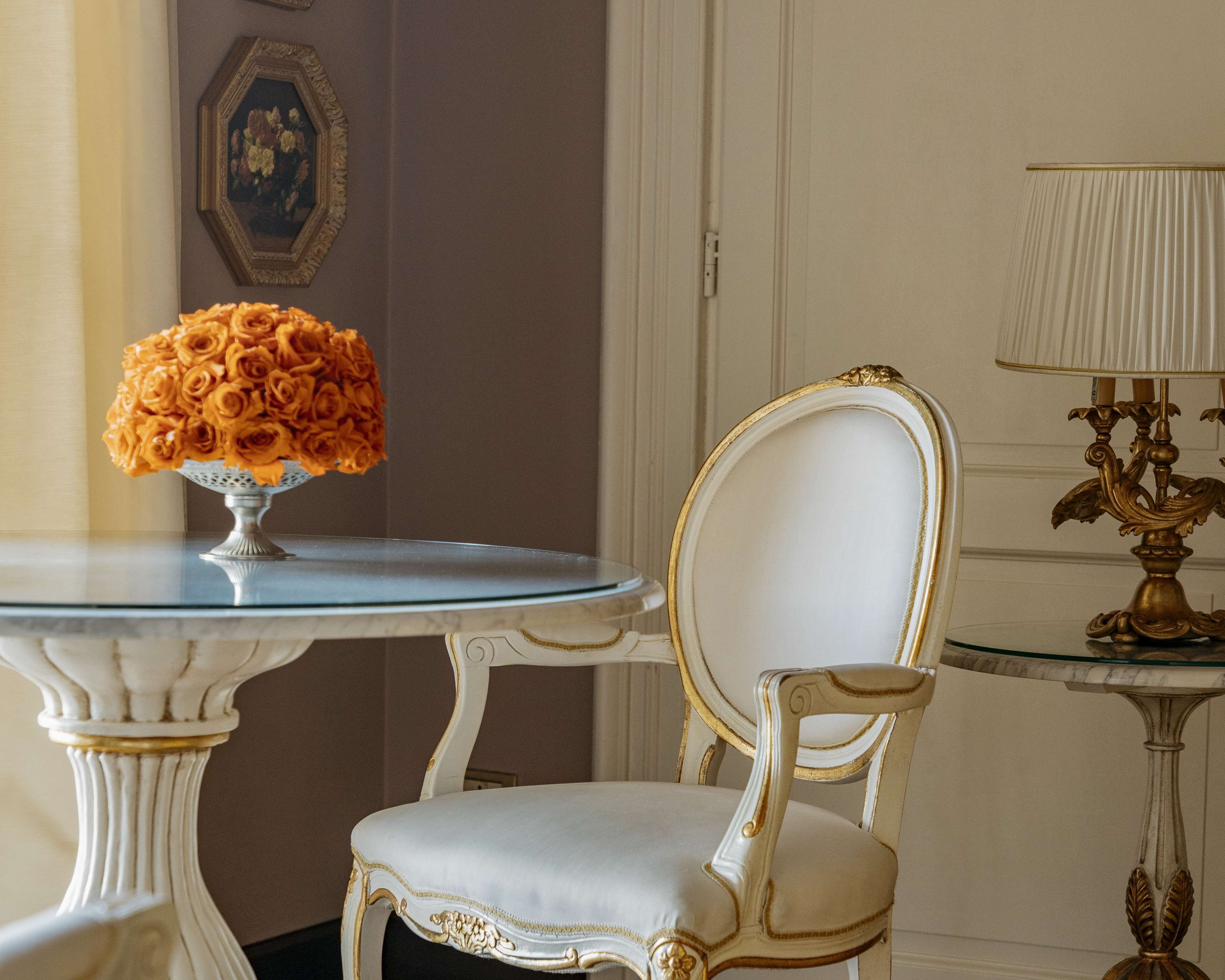 Una habitación con una silla blanca y ornamentada, una mesa de vidrio con un florero y una lámpara de base dorada.