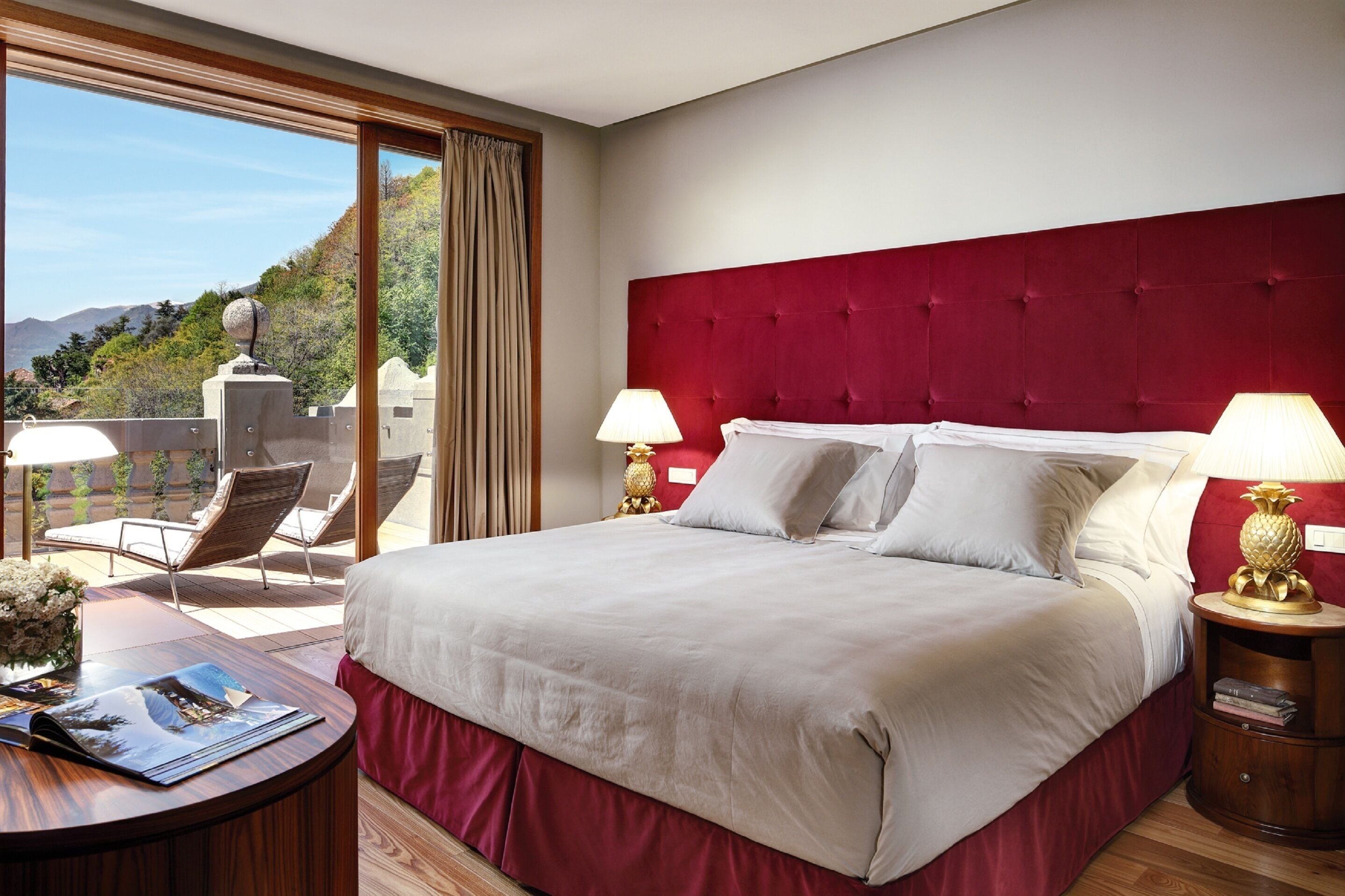 Ein Hotelzimmer mit einem großen Bett, zwei Nachttischlampen, einem Holznachttisch, einem Schreibtisch mit Stuhl und Blick ins Freie.