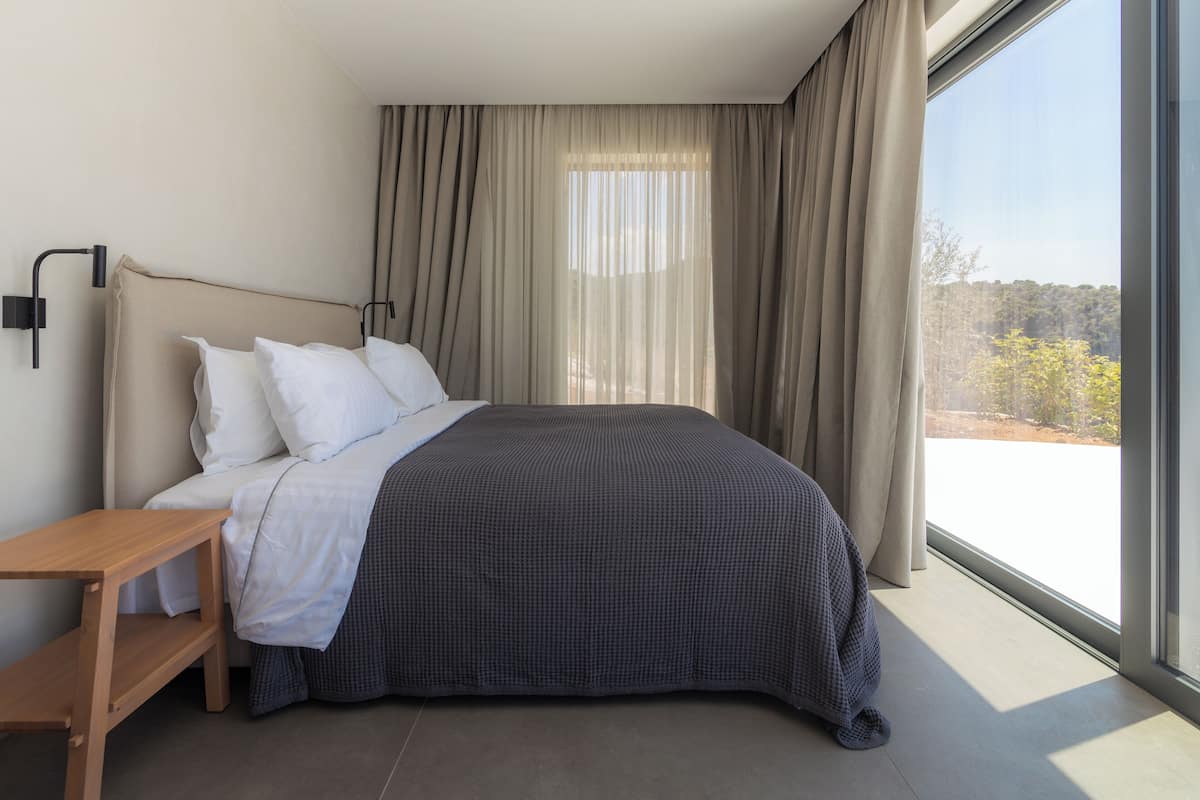 Ett modernt hotellrum med en stor säng, ett träbord vid sängen och utsikt över omgivningarna genom ett stort fönster.