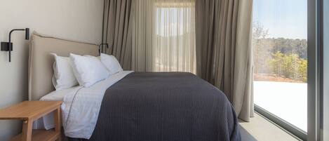 Una habitación de hotel moderna con una cama grande, una mesita de noche de madera y vistas al exterior a través de un ventanal amplio.
