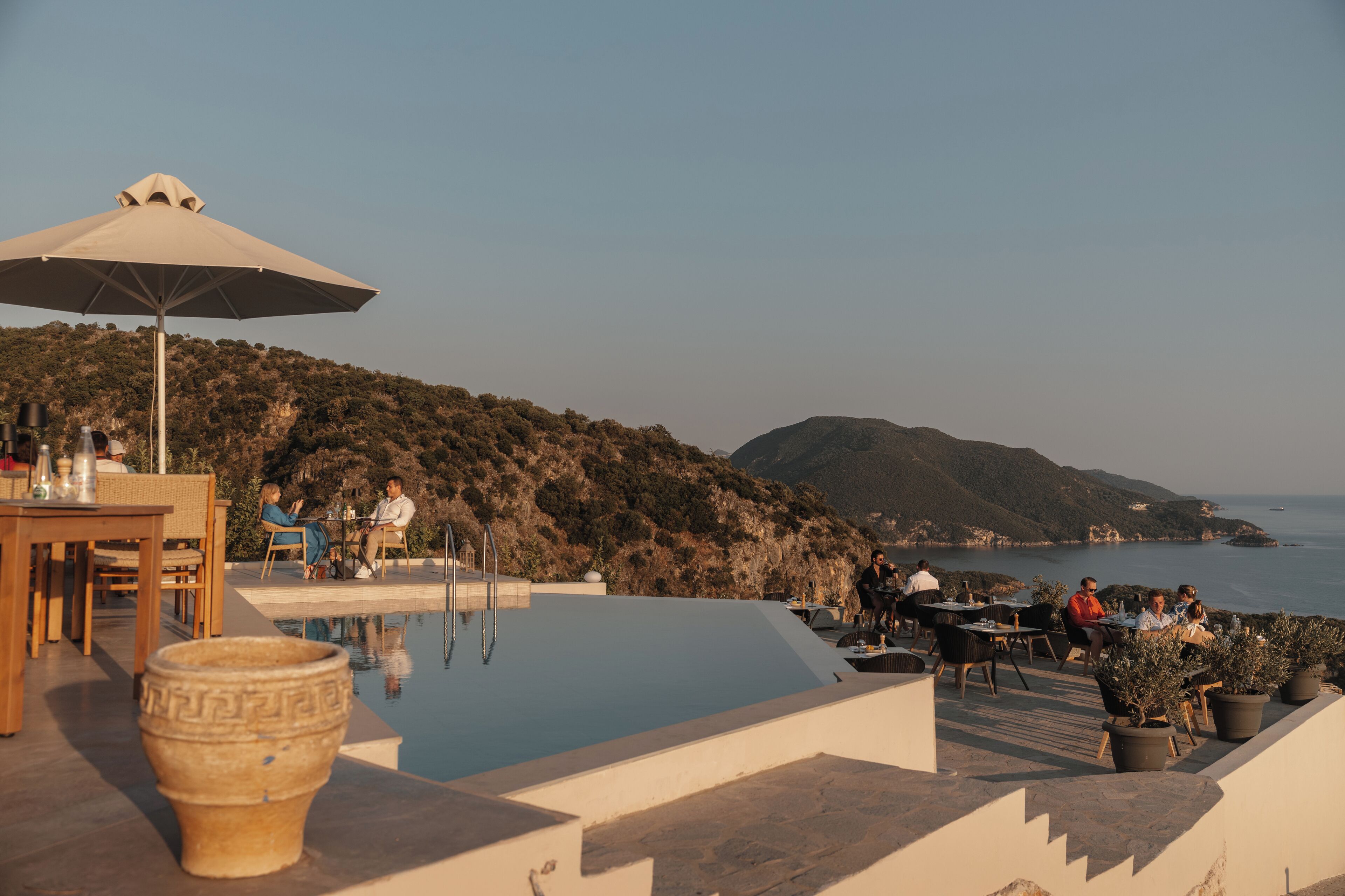 Area piscina esterna con tavoli e sedie, un ombrellone grande e vista sul mare e sulle colline.
