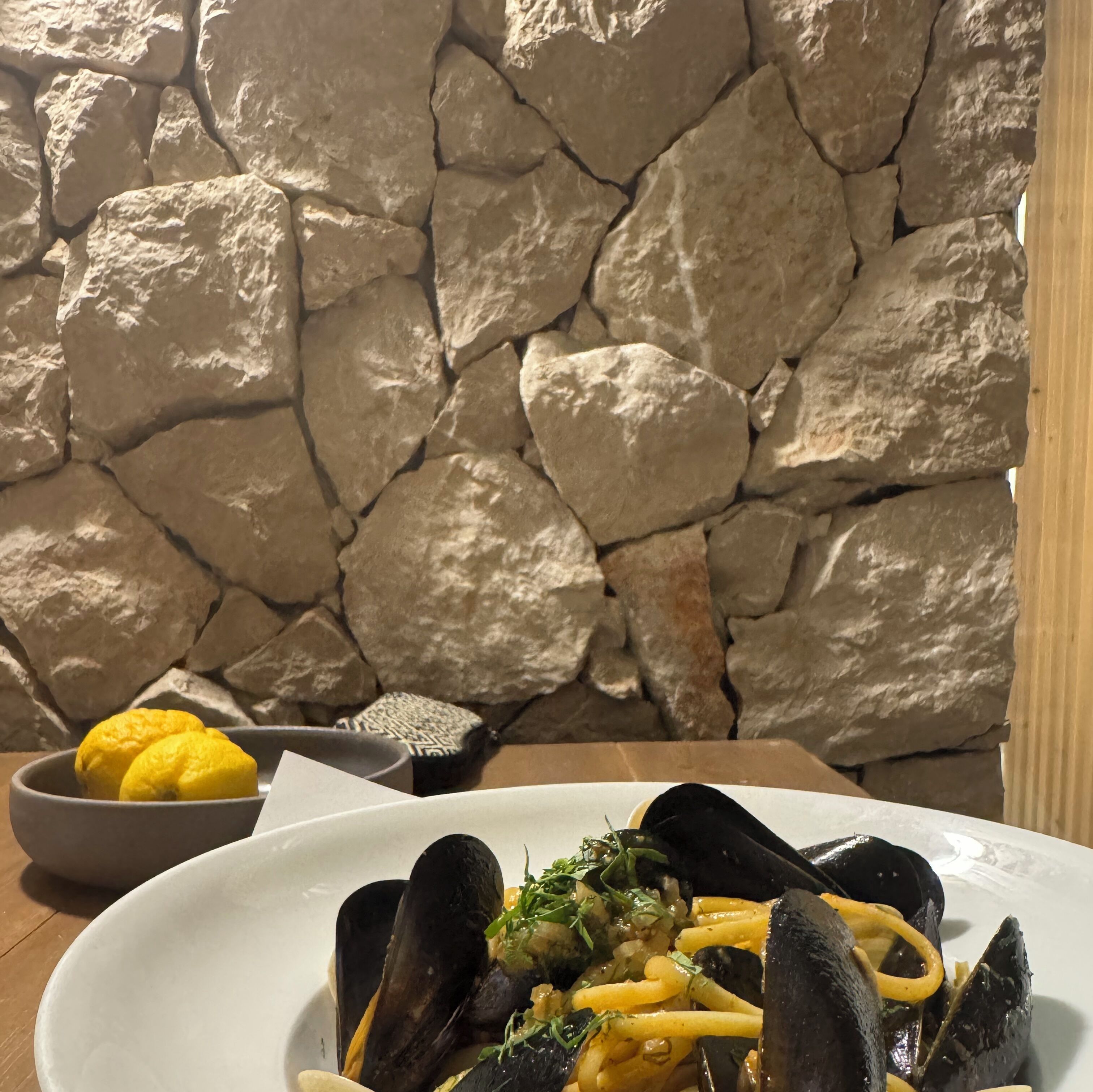 A seafood dish with mussels and pasta, a bowl of lemons, and a patterned cushion on a wooden table against a stone wall.