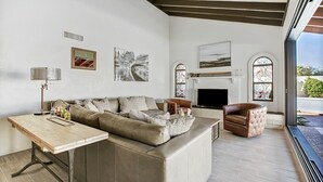 Un amplio salón con un gran sofá, una mesa de centro y una chimenea. Hay dos ventanas con vidrieras decorativas, y obras de arte adornan las paredes.