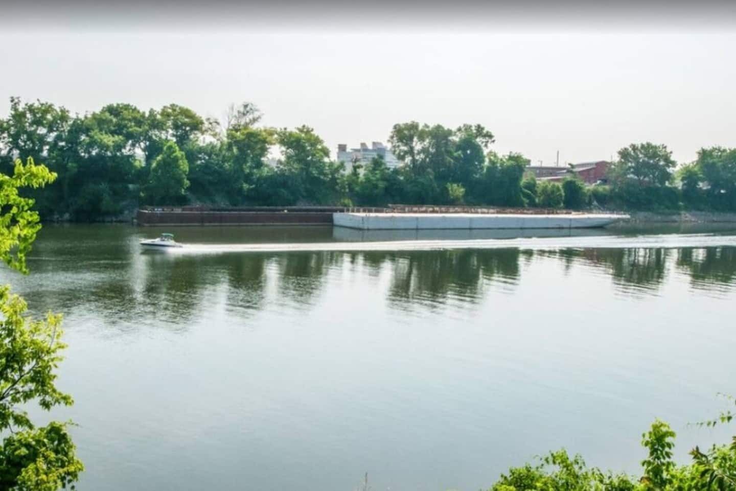 Une rivière paisible, traversée par un bateau, avec des arbres et des bâtiments en arrière-plan.