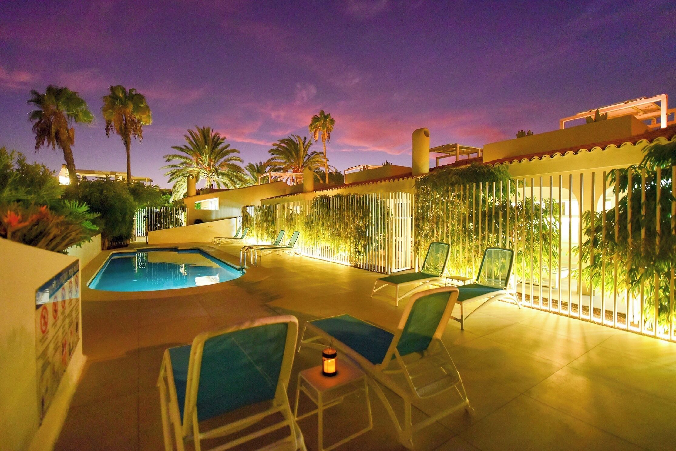 An outdoor pool area with lounge chairs and a small table, surrounded by palm trees and a fence.