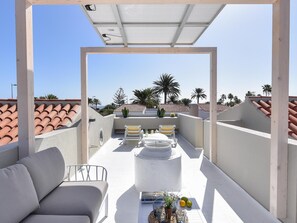 A rooftop terrace with a white ceiling, two cushioned chairs, a small table with drinks, and a potted plant.