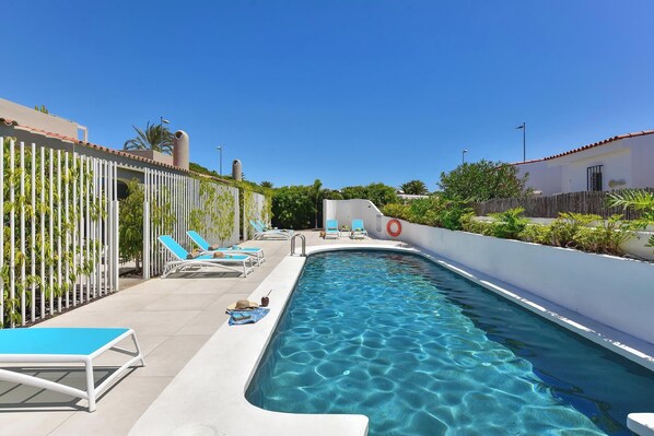 A swimming pool with clear blue water, surrounded by white walls and a fence with greenery. There are lounge chairs and a sunshade on the poolside.