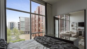 Une chambre d’hôtel moderne avec une grande fenêtre, un lit recouvert d’une couette noire et blanche, une table basse en verre, un canapé et un téléviseur à écran plat.
