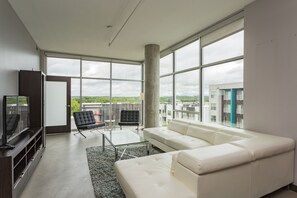 Una sala de estar moderna con un sofá blanco, una mesa de centro de vidrio y una silla negra. Una ventana grande ofrece vistas a edificios y zonas verdes.